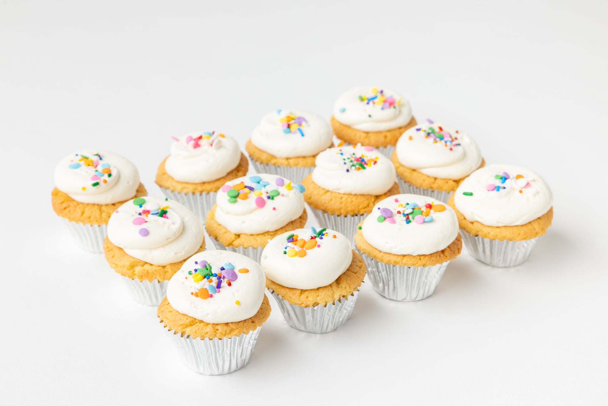 Row of vanilla cupcakes with white frosting and colorful sprinkles on a light background